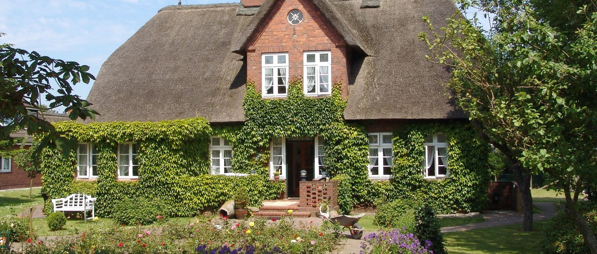 Großes altes Landhaus mit Reetdach, roter Klinkerfassade und grünem Garten – Symbolbild für eine Immobilie, die man vererben kann.