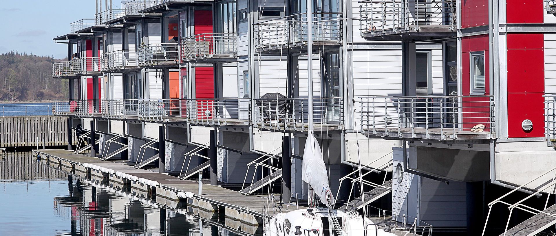 Moderne Reihenhäuser direkt am Wasser mit Bootssteg und Segelboot – Symbolbild für teure Immobilien in begehrter Lage.