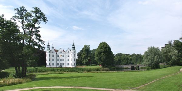 Helles Schlossgebäude mit Türmen am Rande eines großen Rasens, umgeben von Bäumen und Wasserlauf