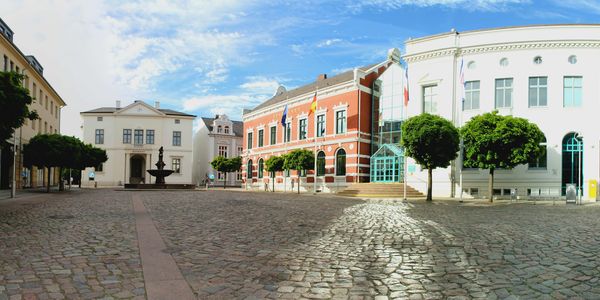 Historisches Rathausensemble auf dem Marktplatz von Bad Oldesloe mit Brunnen, Kopfsteinpflaster und gepflegten Bäumen bei blauem Himmel