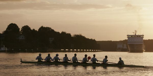 Ruderteam mit neun Personen auf einem ruhigen Gewässer bei Sonnenuntergang. Im Hintergrund fahren große Schiffe vorbei.