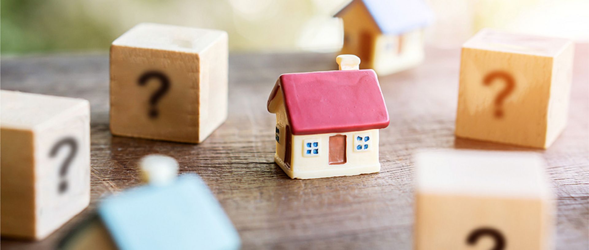 Ein kleines Spielzeughaus mit rotem Dach steht auf einem Holztisch, umgeben von Würfeln mit Fragezeichen und weiteren kleinen Häusern.