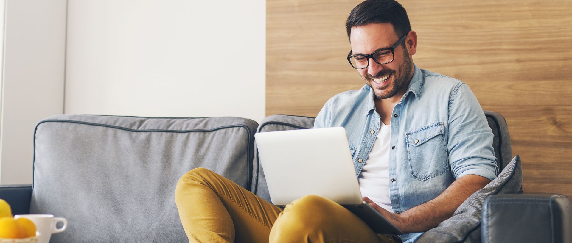 Ein Mann sitzt entspannt auf einem grauen Sofa und arbeitet oder surft mit einem Laptop. Er trägt eine Brille, ein Jeanshemd und senffarbene Hose und lächelt dabei.