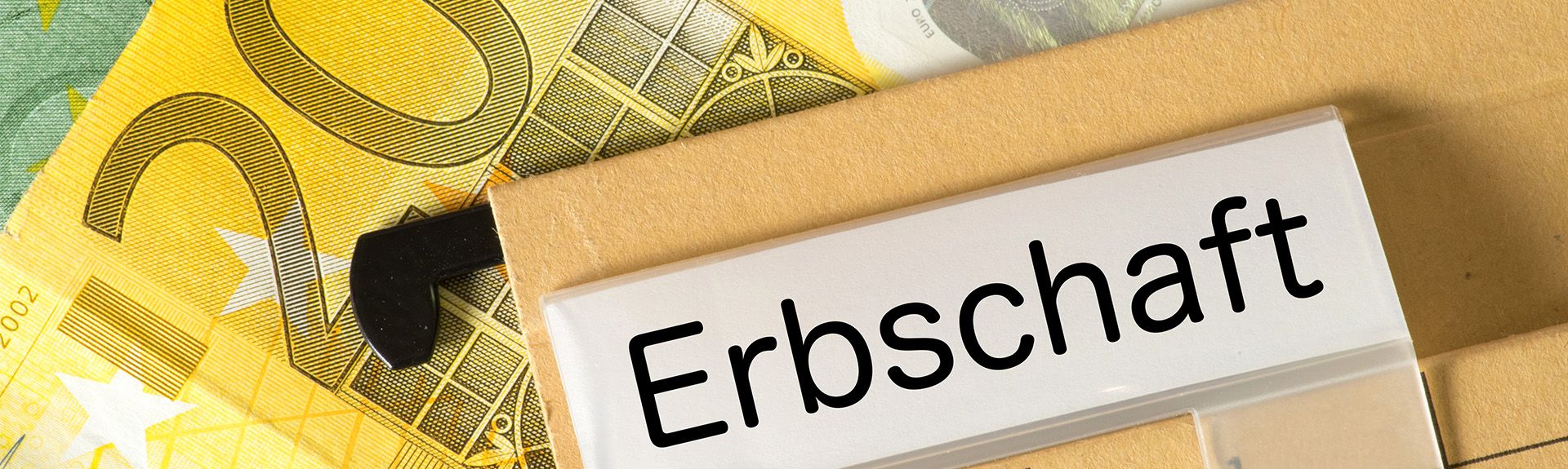 Ein Aktenordner mit dem beschrifteten Reiter „Erbschaft“ liegt auf mehreren Euro-Geldscheinen. Im Vordergrund ist der schwarze Griff der Ordnerrückseite zu sehen.