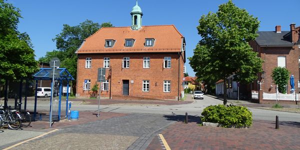 Ein Platz in der Innenstadt von Schwarzenbek mit einem roten Backsteingebäude und kleinem Turm. Im Vordergrund befinden sich eine Bushaltestelle, Fahrräder und ein Baum mit Blumenbeet. Der Himmel ist wolkenlos blau.